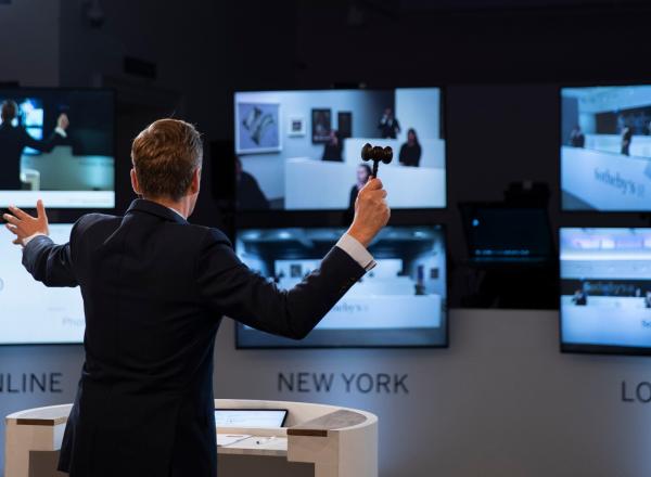 an auctioneer with a gavel stands in front of three large computer screens